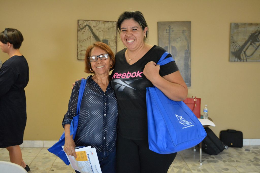 Two women pose together for a photo. They are both smiling and holding goodie bags given out by Community Vitals Signs to all in attendance.